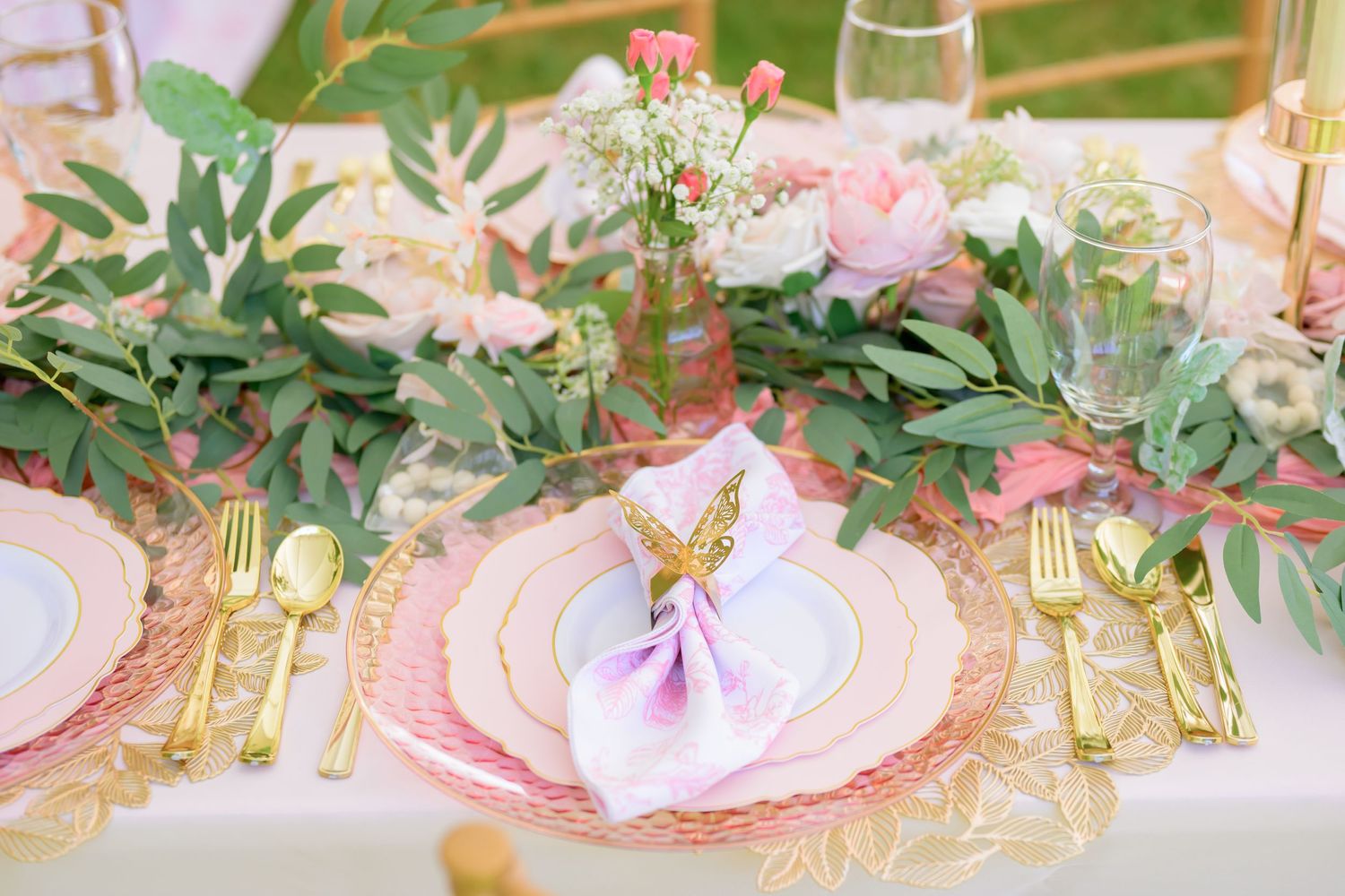 Romantic table setting with pink plates, gold cutlery, and fresh greenery centerpiece.