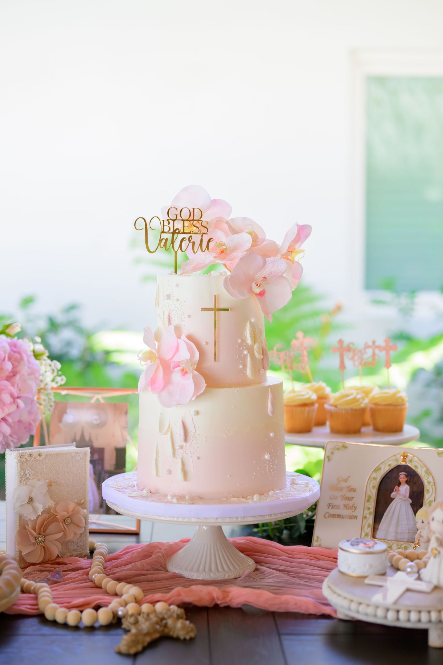 Pink and white two-tier First Communion cake with gold cross topper and religious decorations.