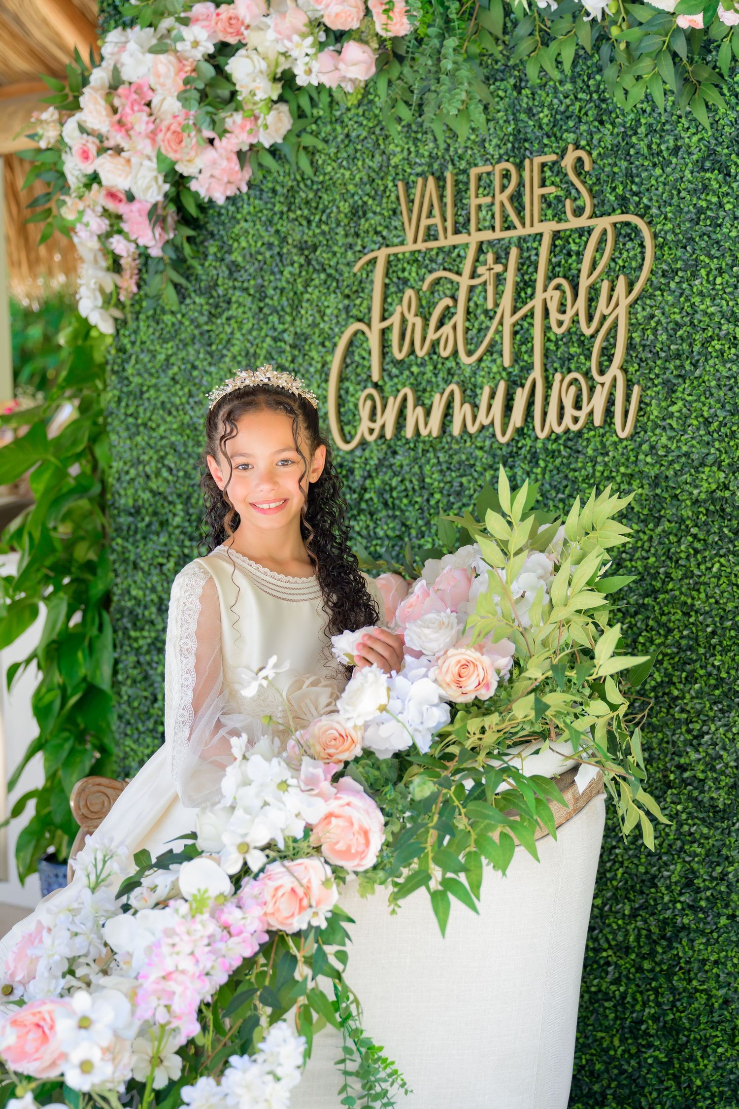Elegant communion celebration backdrop with green hedge wall, gold lettering, and pink floral decorations.