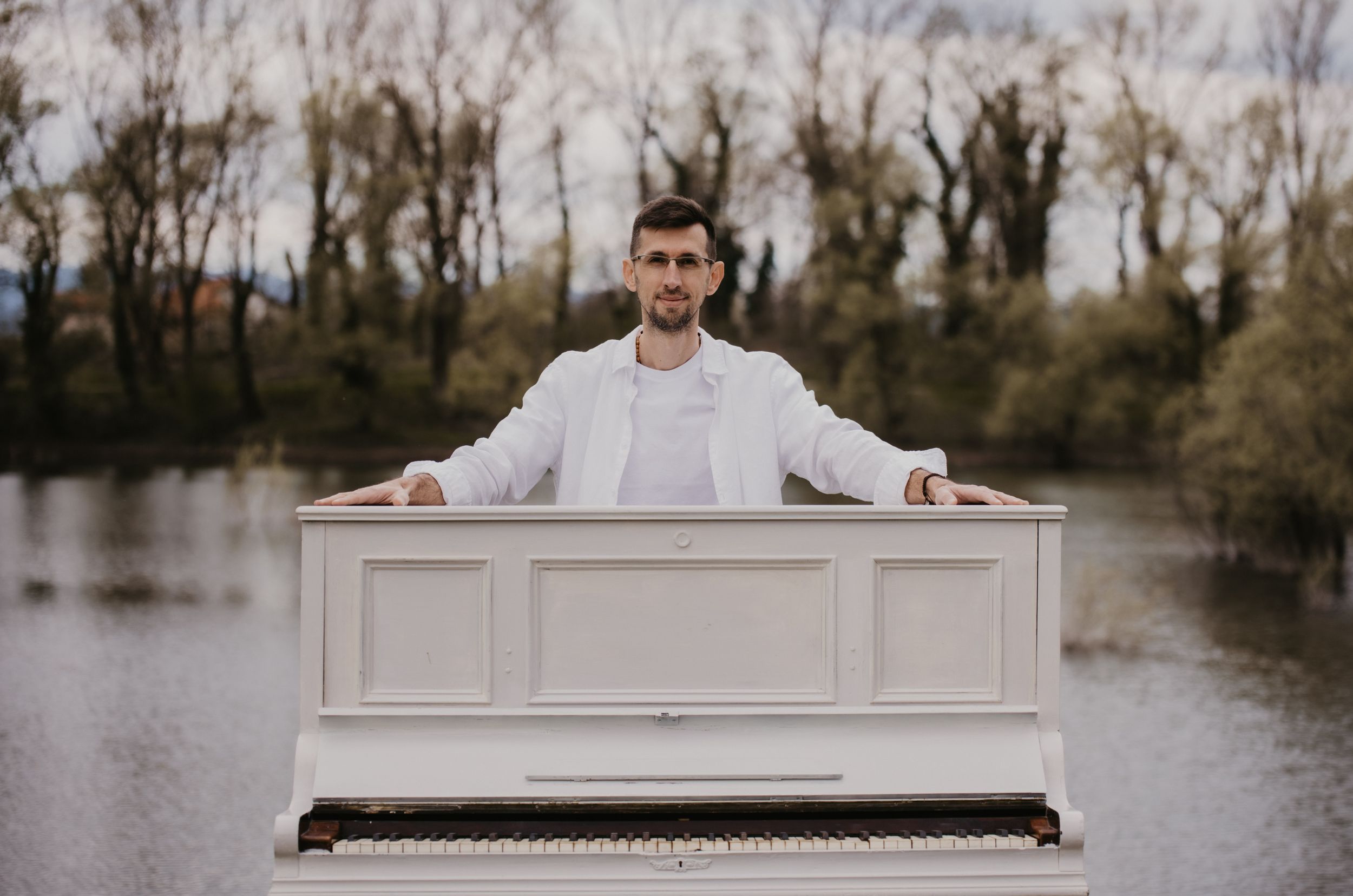 A white piano sits beside a serene lake with bare trees in the background.