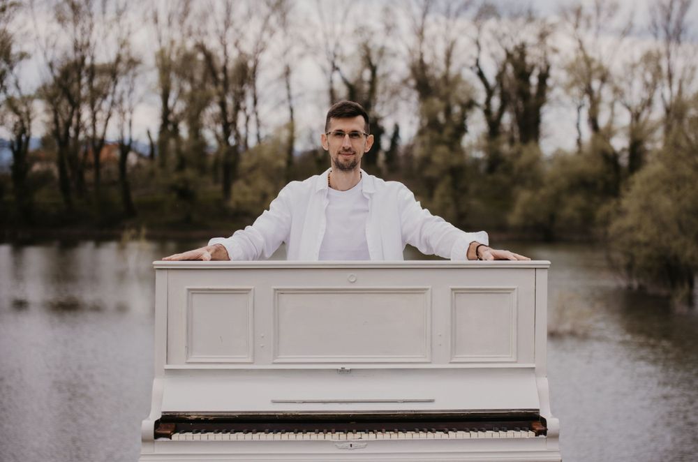 A white piano sits beside a serene lake with bare trees in the background.