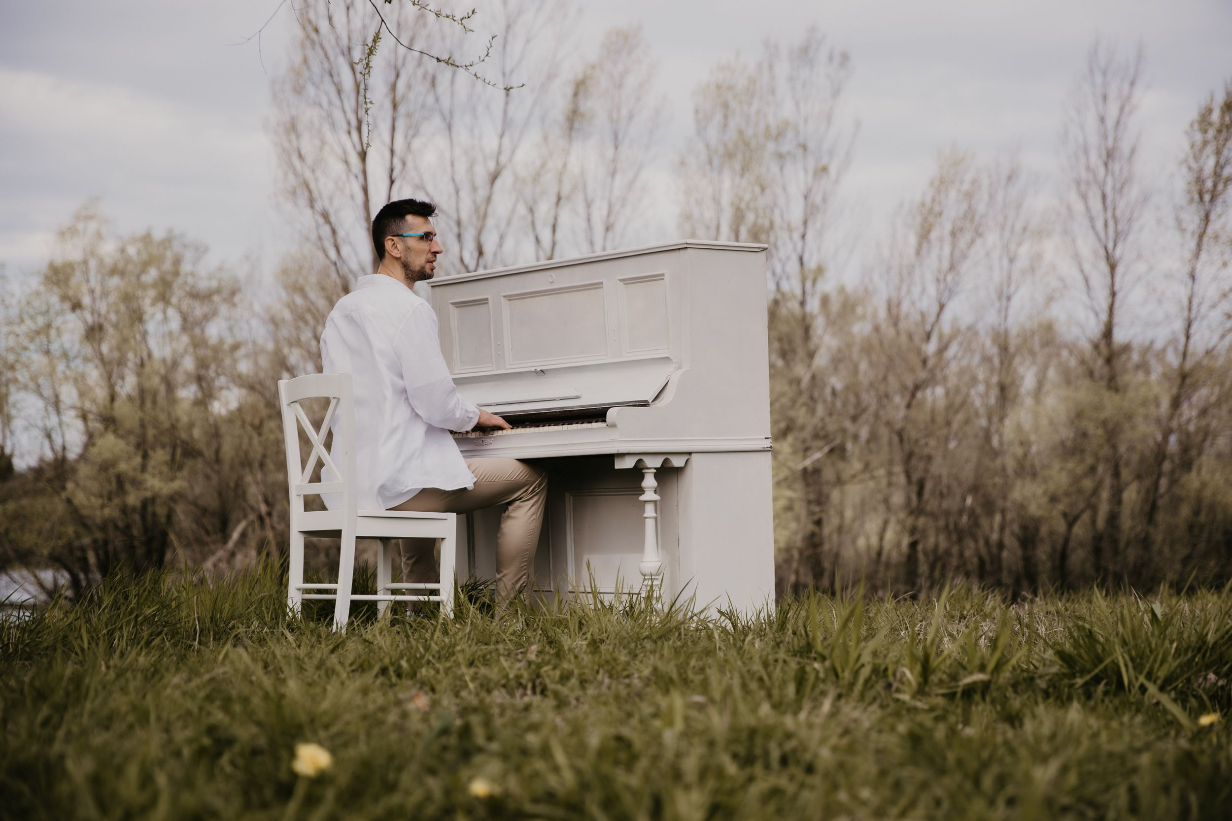 White upright piano placed in grassy field with bare trees in background.