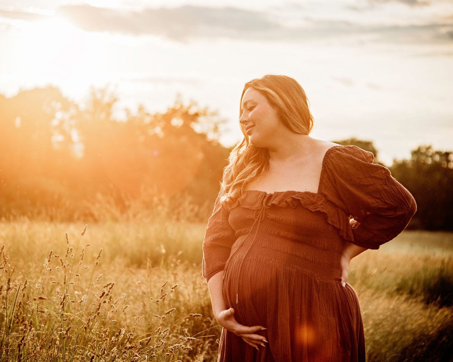 sweetly smiling mama posing with her baby bump in a golden lighted meadow for outdoor maternity photoshoot