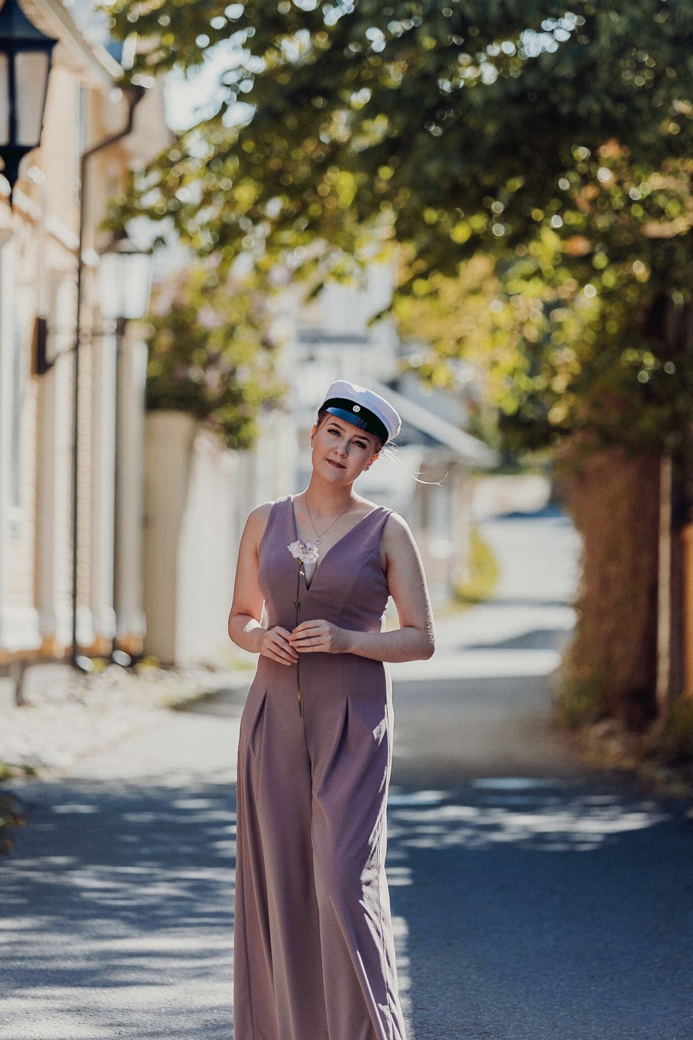 Person wearing a flowing mauve dress standing on a sunny street with trees in background.
