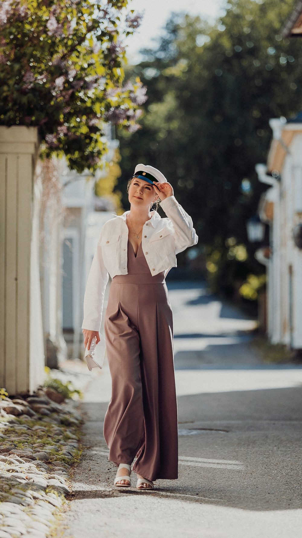 Figure in a mauve dress and white shawl walking along a tree-lined street on a sunny day.