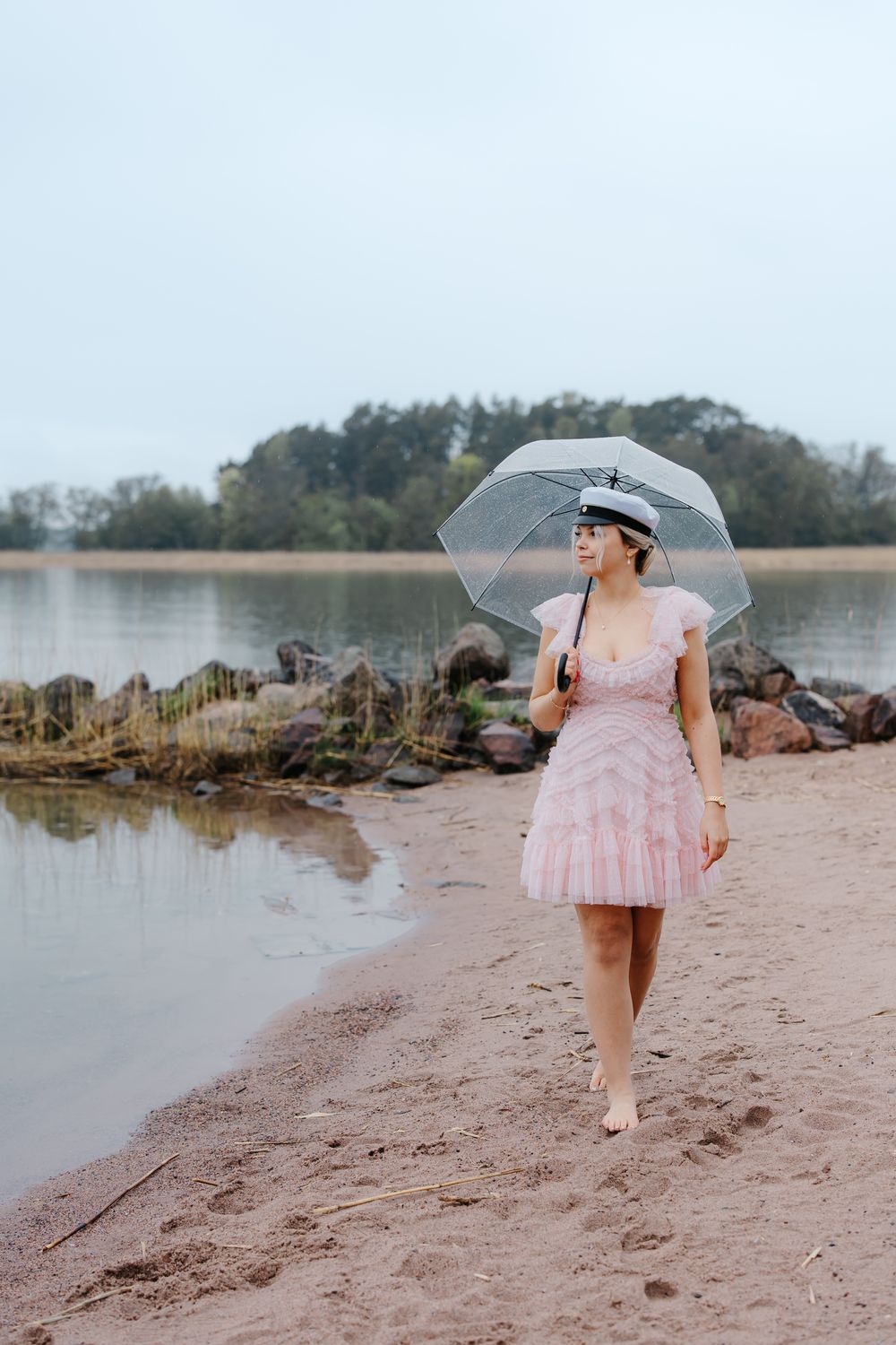 Woman in pink dress and white cap walks barefoot on sandy beach holding clear umbrella in light rain.