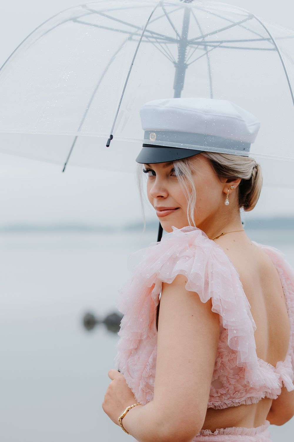 Woman in pink ruffled dress and white captain hat holds clear umbrella near misty lake.