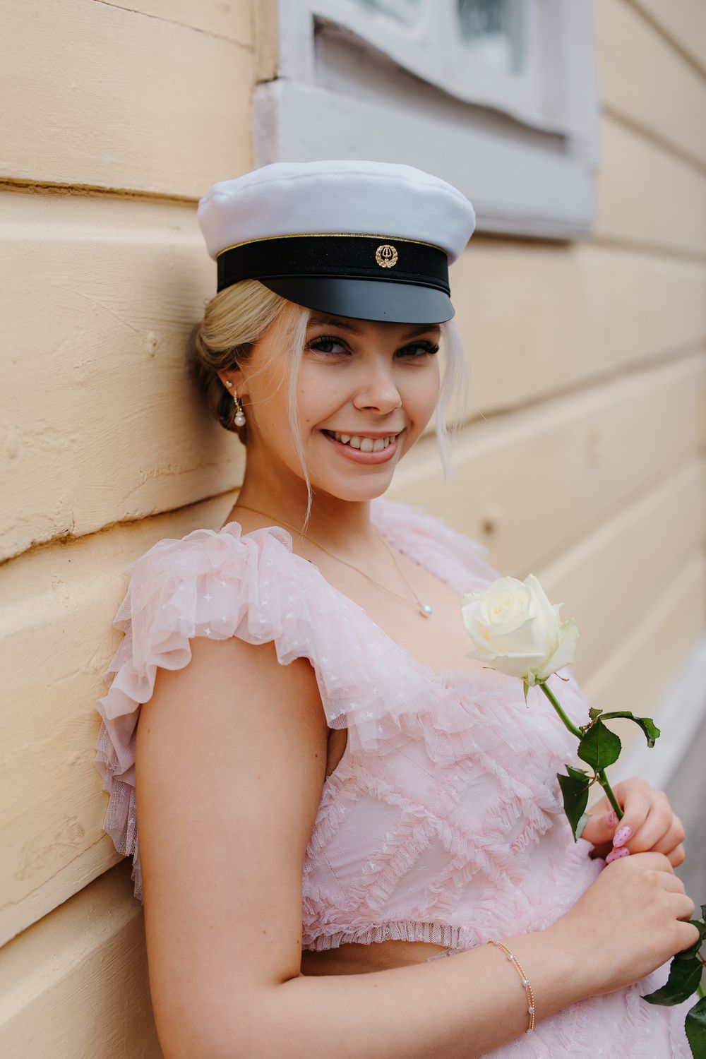 Smiling young woman in pink dress holding white rose, wearing white and black captain hat, leaning against yellow wall.