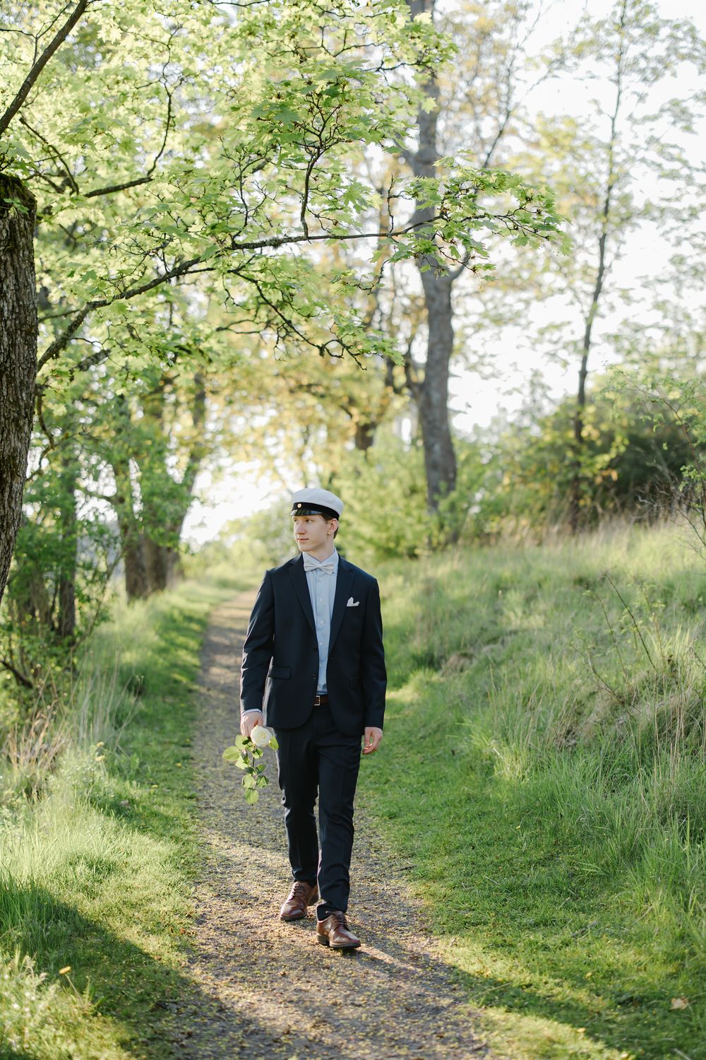 Groom in dark suit and flat cap walks a scenic tree-lined path holding a small bouquet.