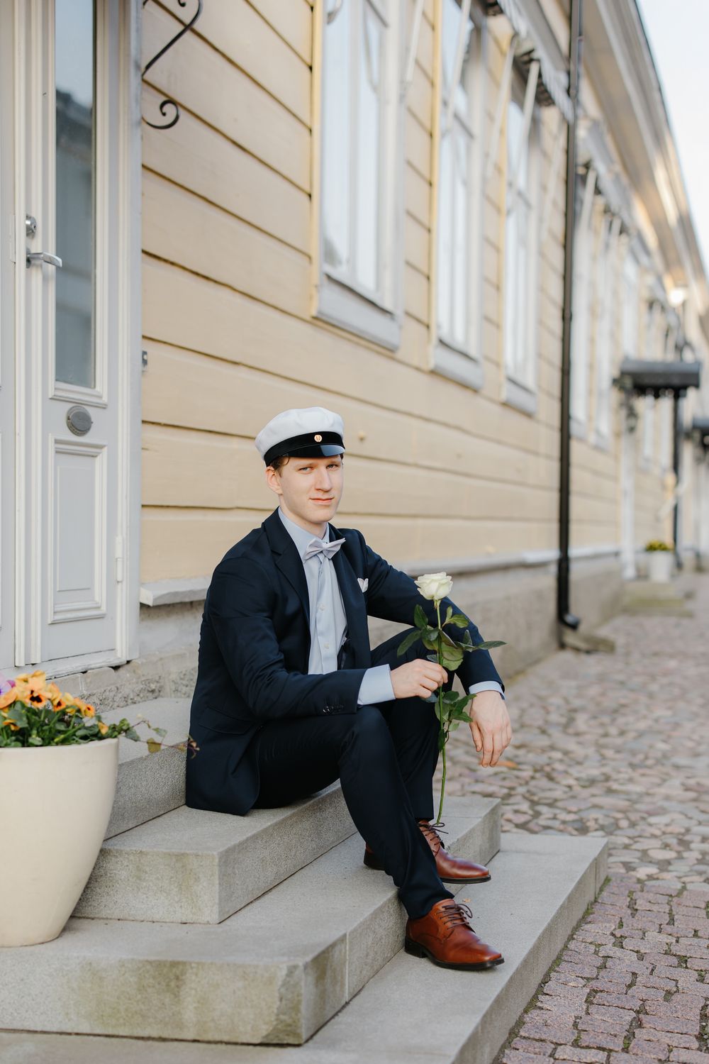 Young man in navy suit and white cap sits on steps holding a white rose outside a historic building.
