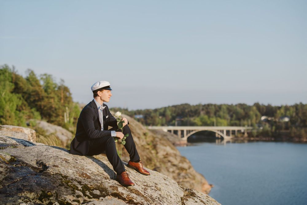 A well-dressed groom in a dark suit and flat cap sits on rocky cliffs holding flowers, with a bridge and blue water behind him.
