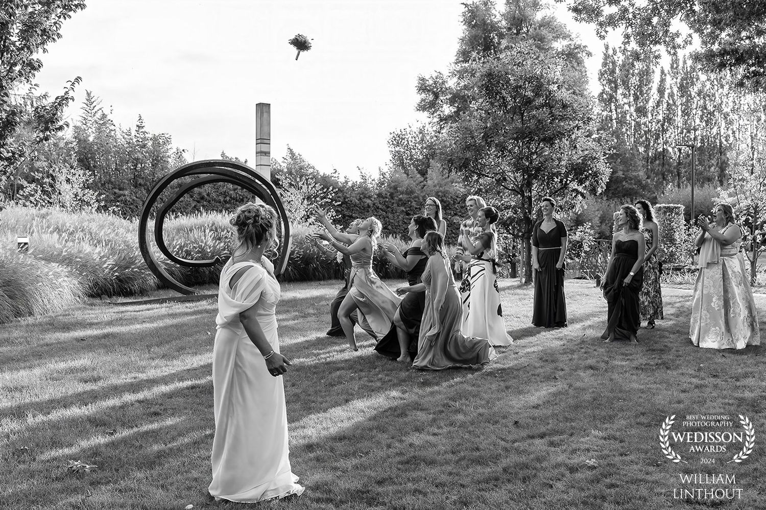 Wedding party in light dresses throws petals outdoors in black and white photograph.