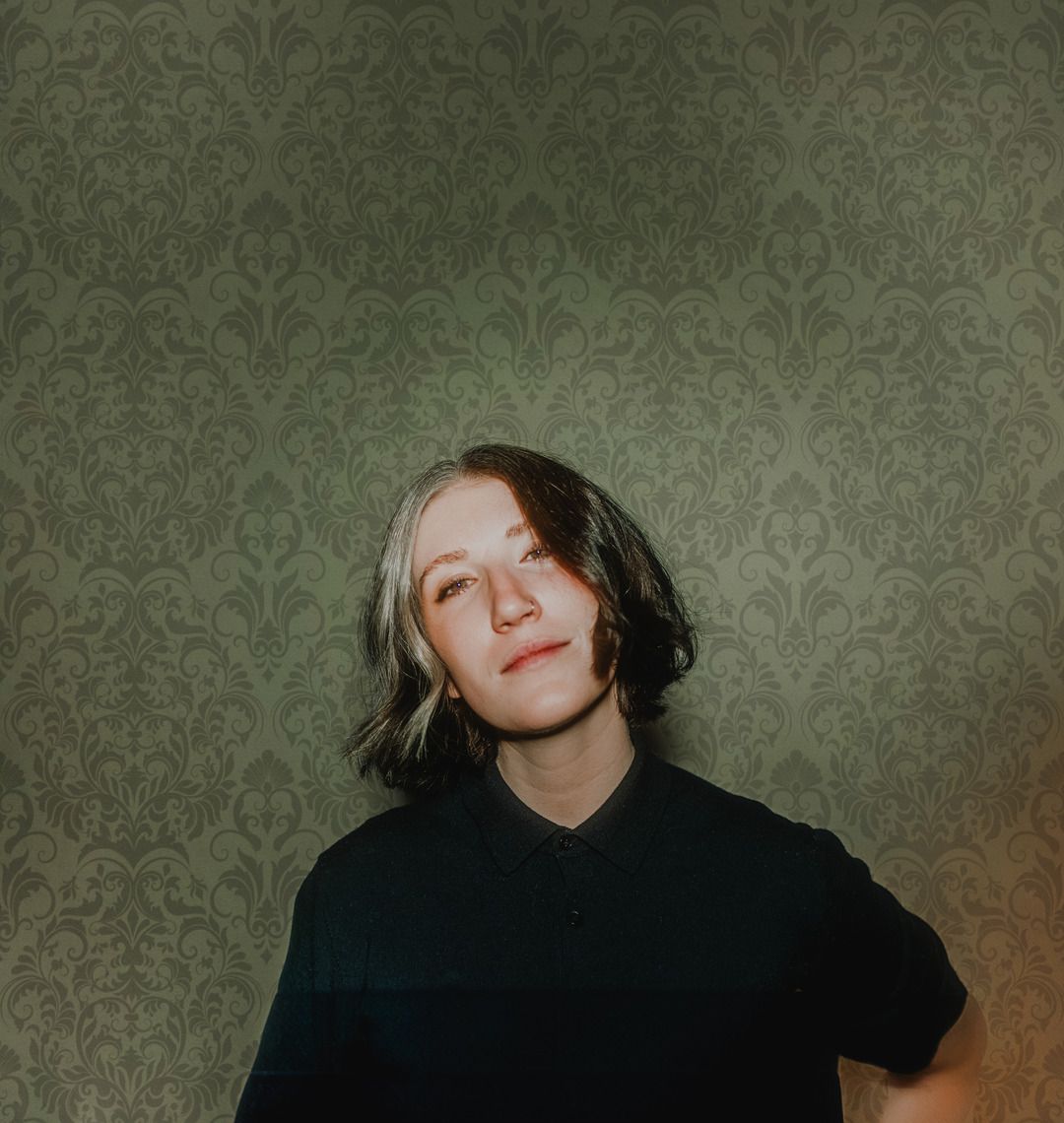 A moody portrait against vintage wallpaper with dramatic lighting and shadows.