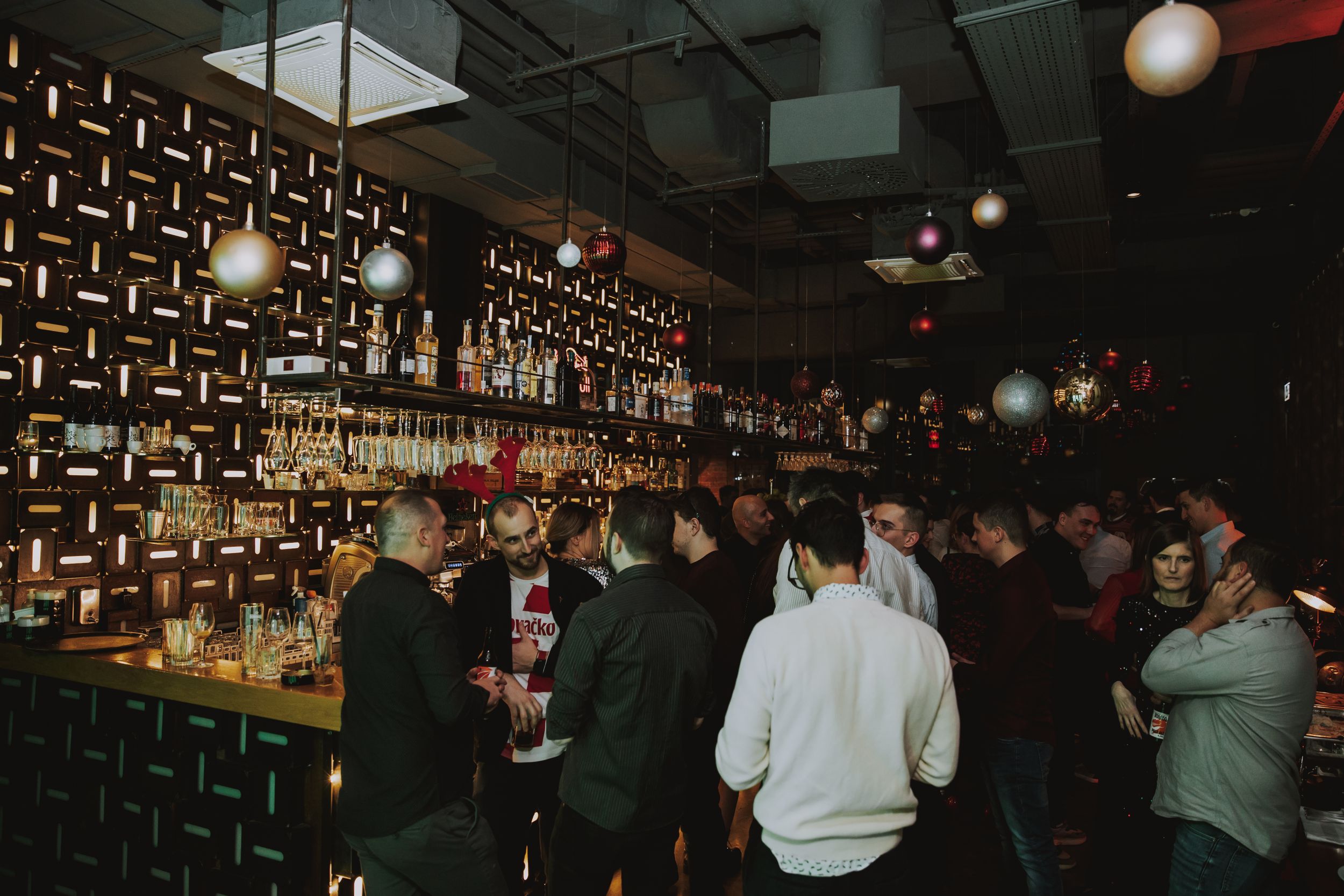 Crowded bar scene with patrons gathered around well-stocked shelves of bottles.