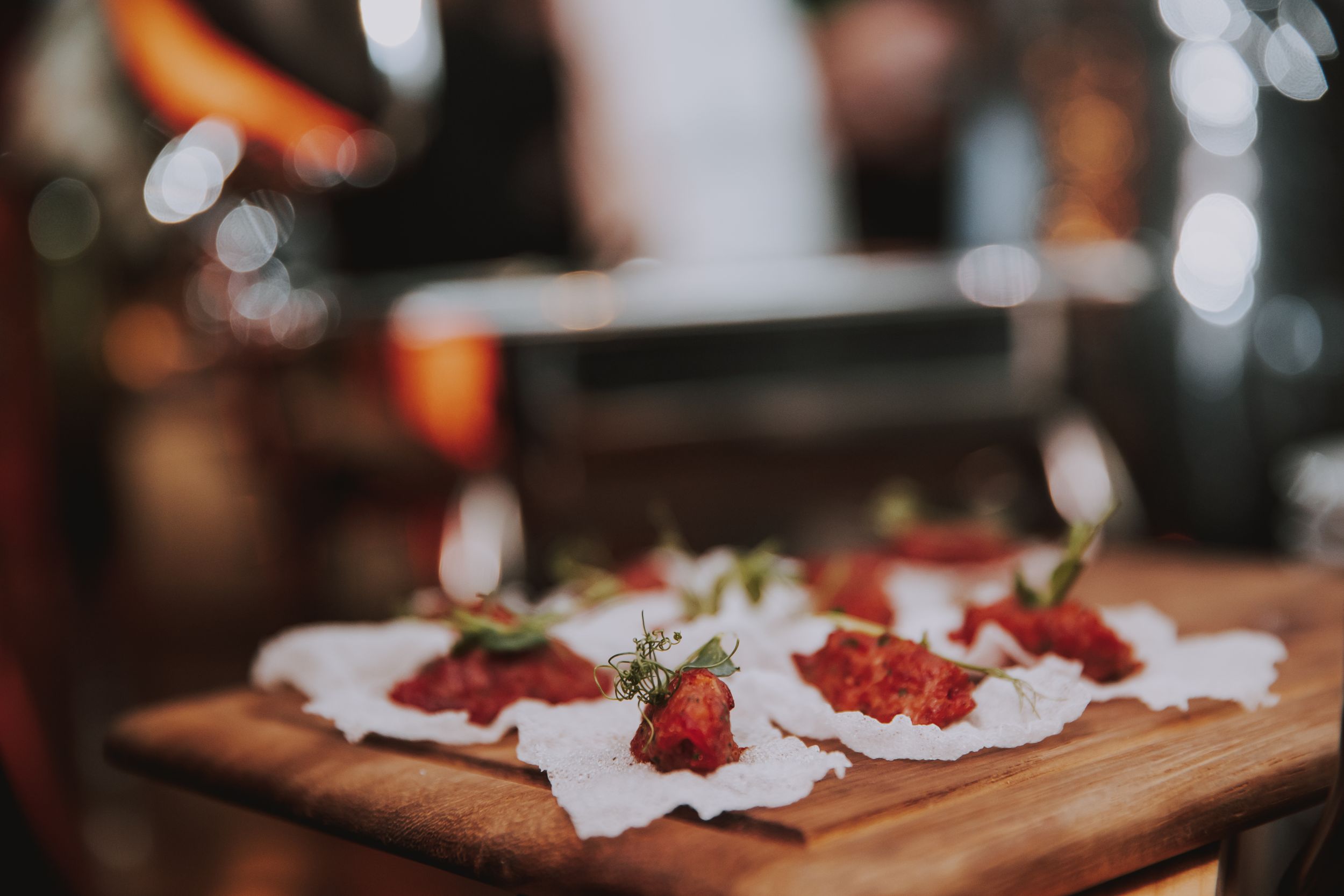 Small appetizers with garnish arranged on white paper atop a wooden serving board in dim lighting.