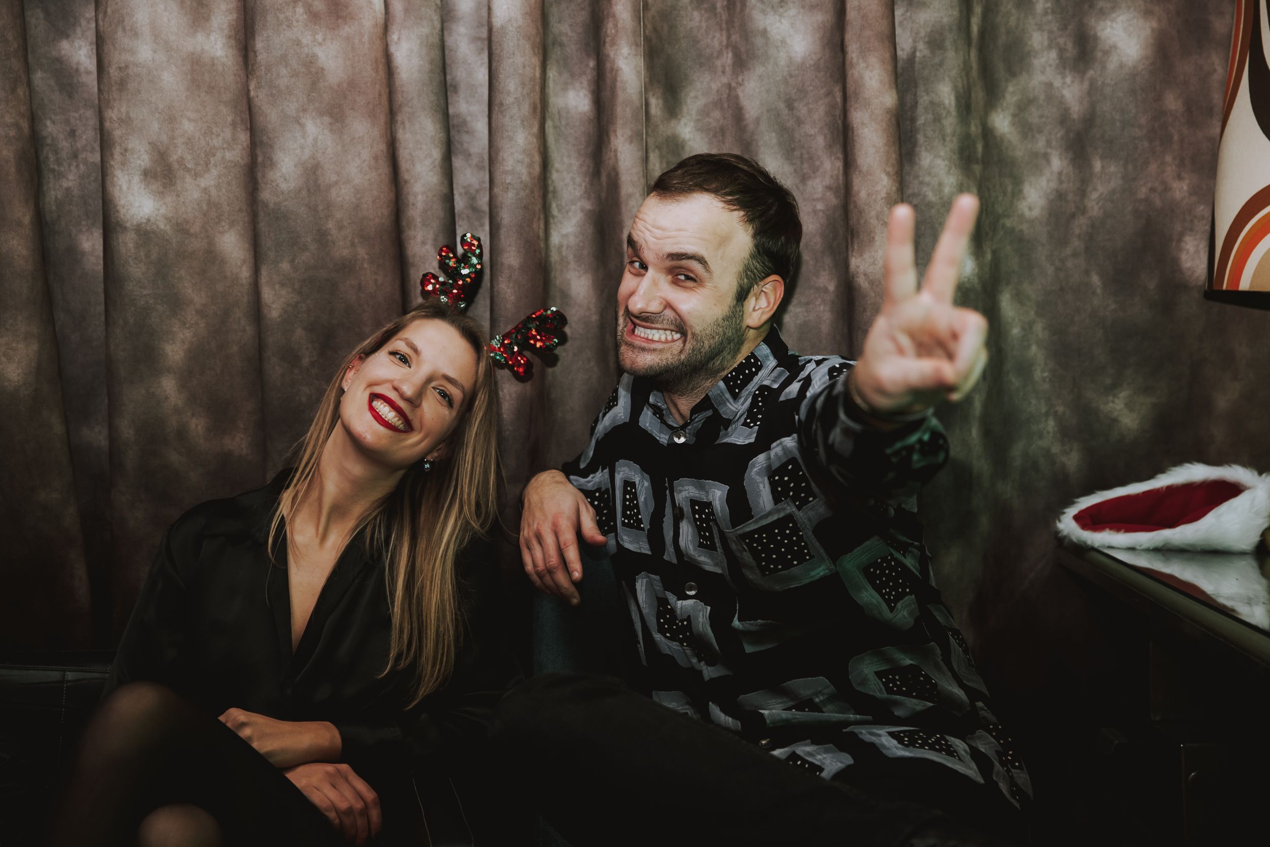 Two friends sharing a joyful moment on a couch at a party, making peace signs and laughing.