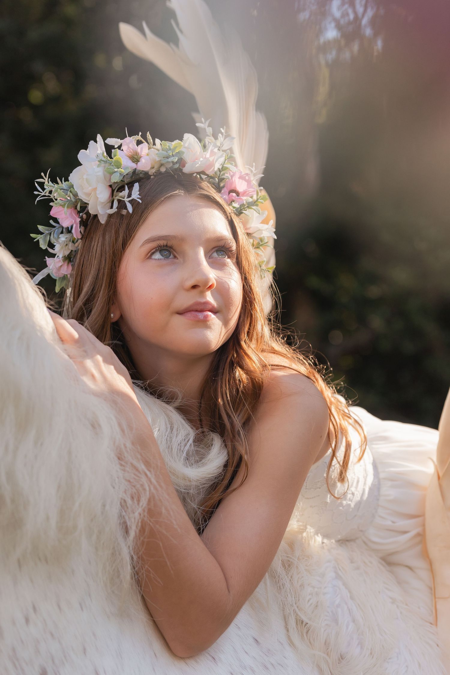Child in white dress with floral crown poses ethereally in golden sunlight.