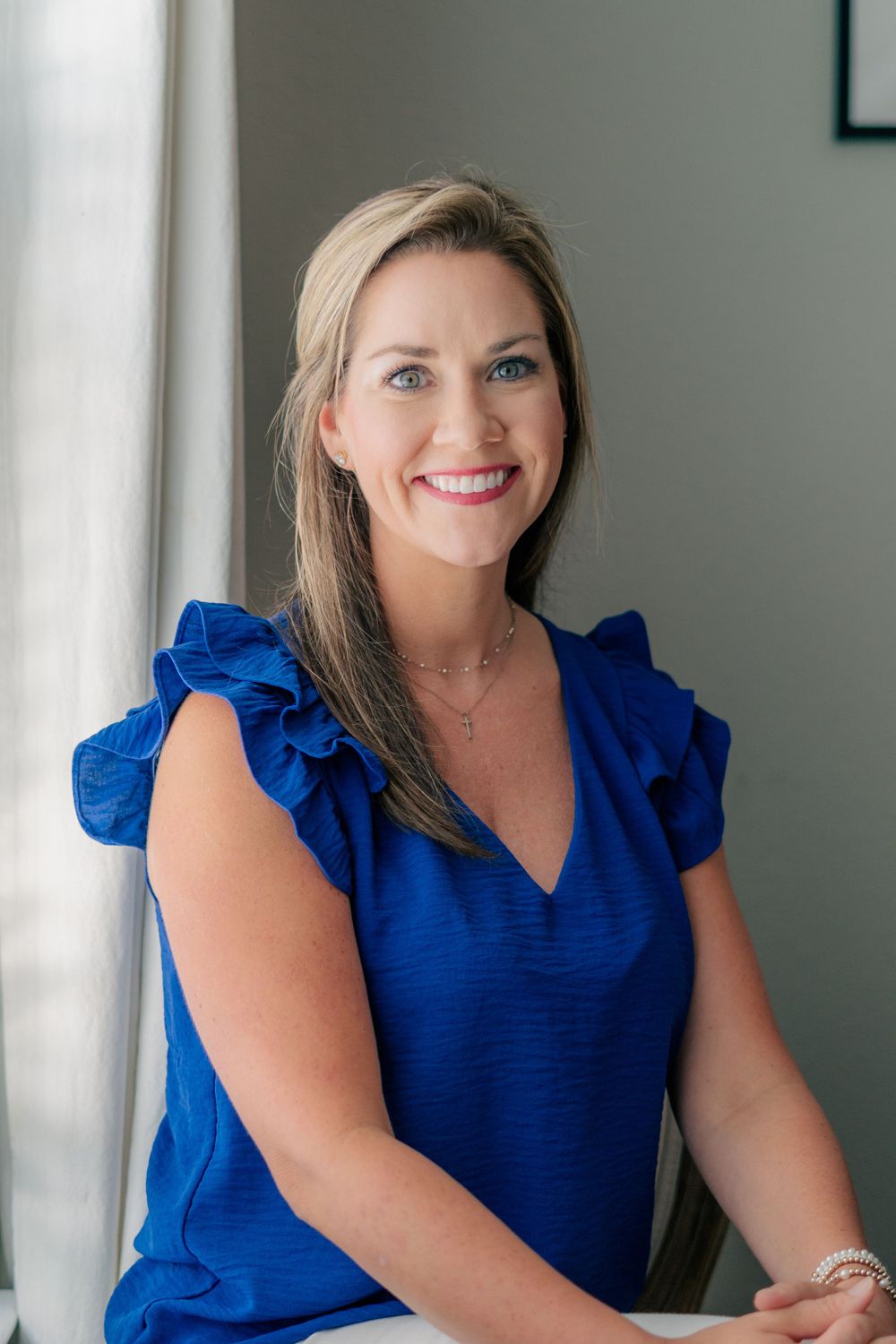 Professional headshot of woman wearing royal blue ruffled blouse against light wall.