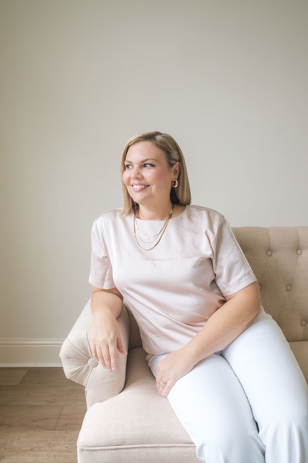 Professional headshot of person in white t-shirt sitting comfortably on beige chair against neutral wall.