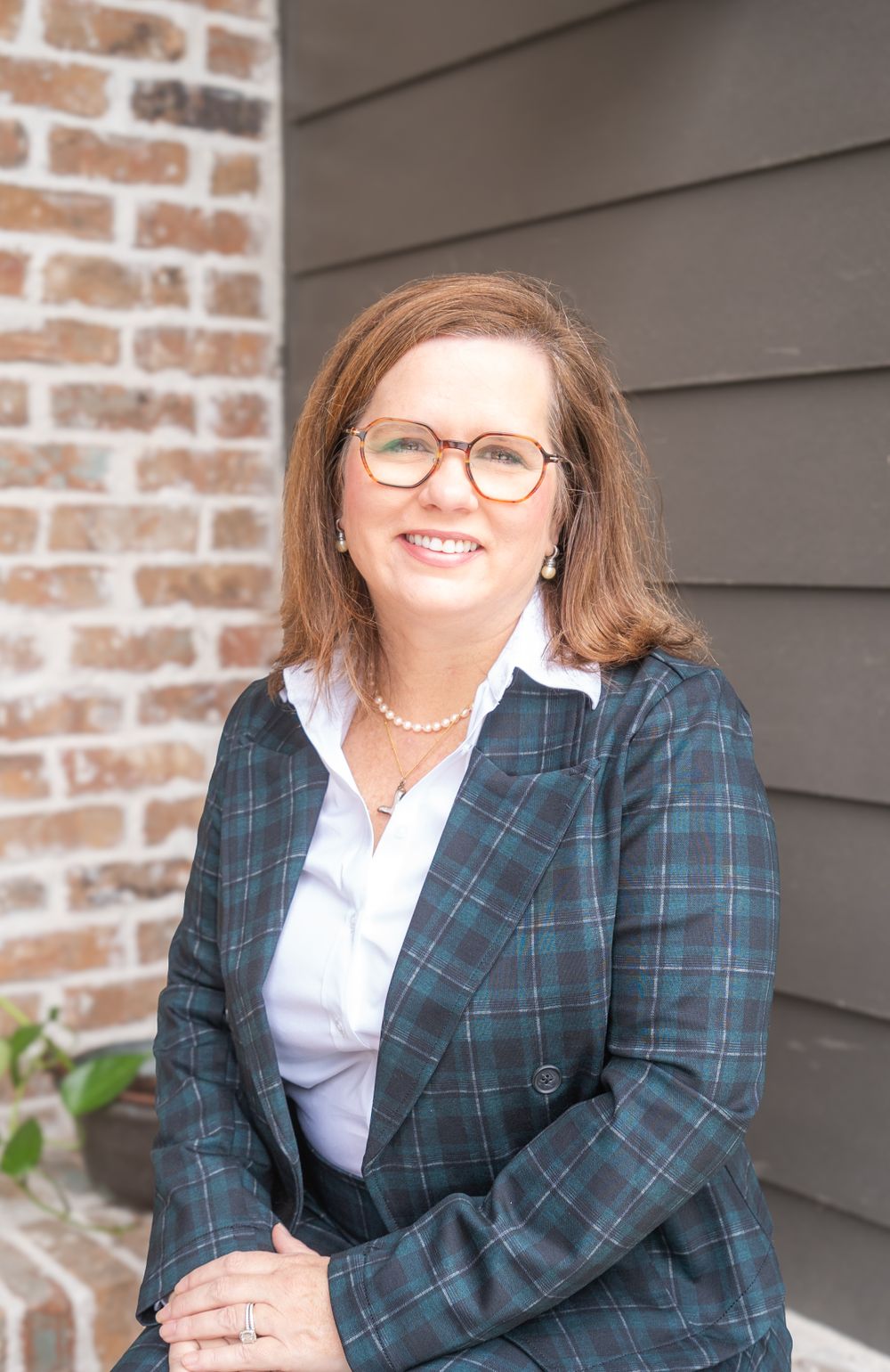 Professional portrait of person in plaid blazer and glasses against brick wall exterior.