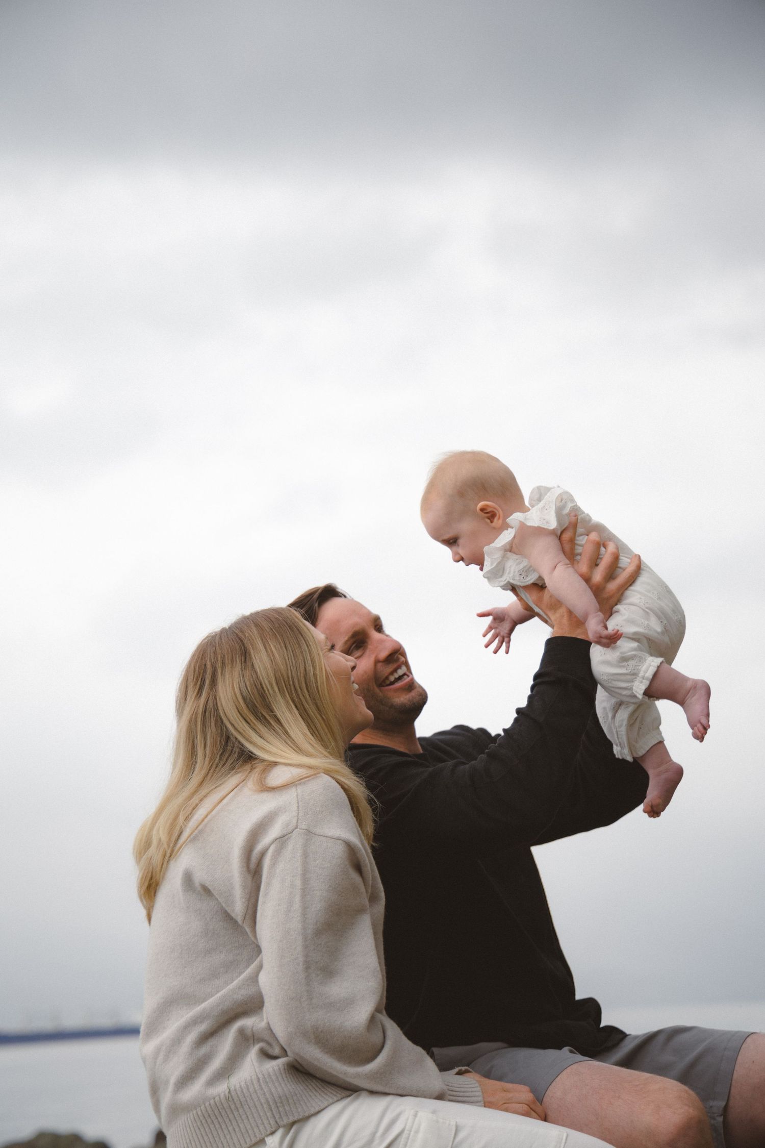 Couple holding up their baby into the air on a cloudy rainy day at the beach in Vancouver, BC sitting on a log.