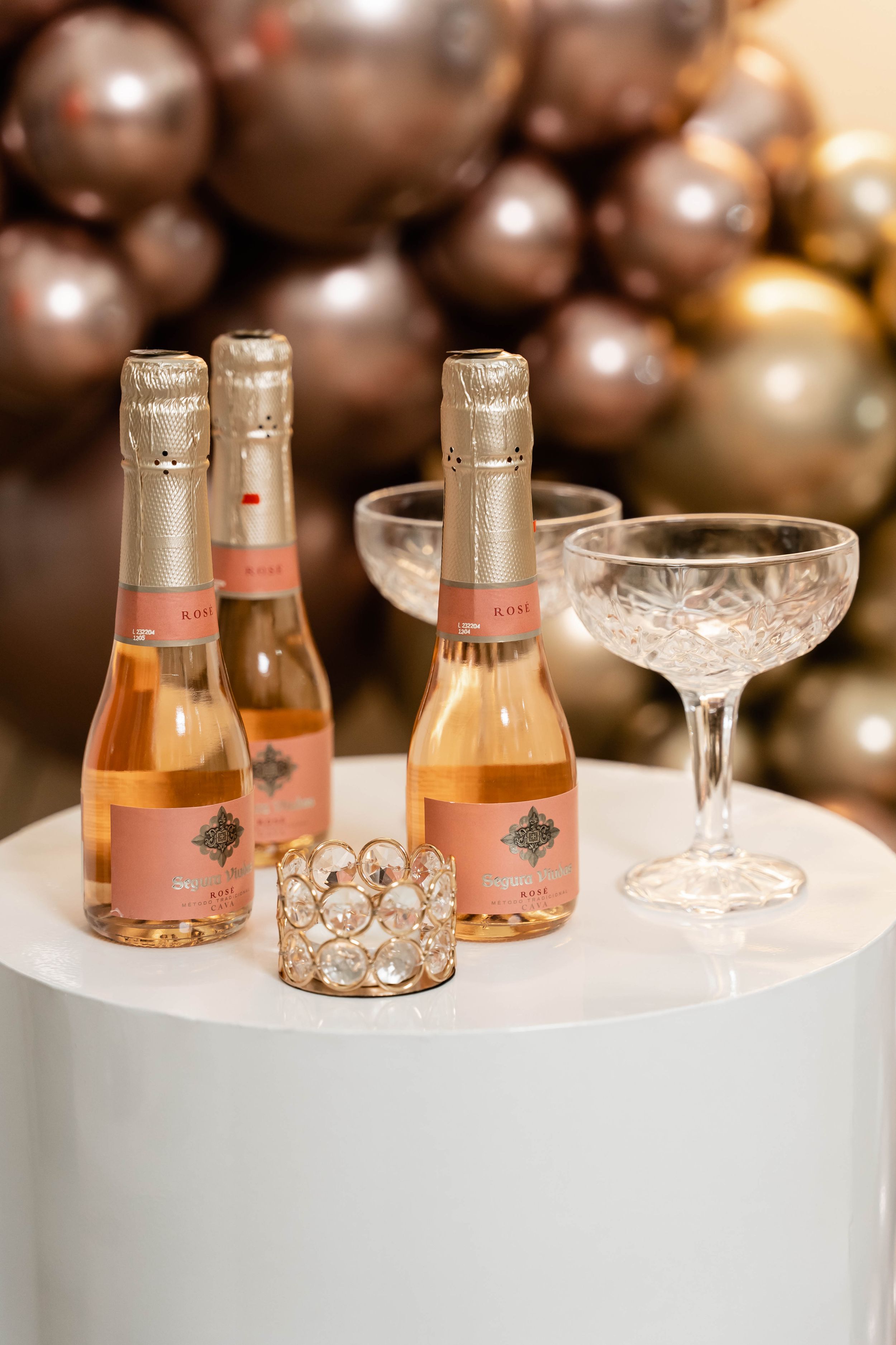 Rose gold champagne bottles and vintage crystal glasses arranged on white pedestal with metallic decorations.