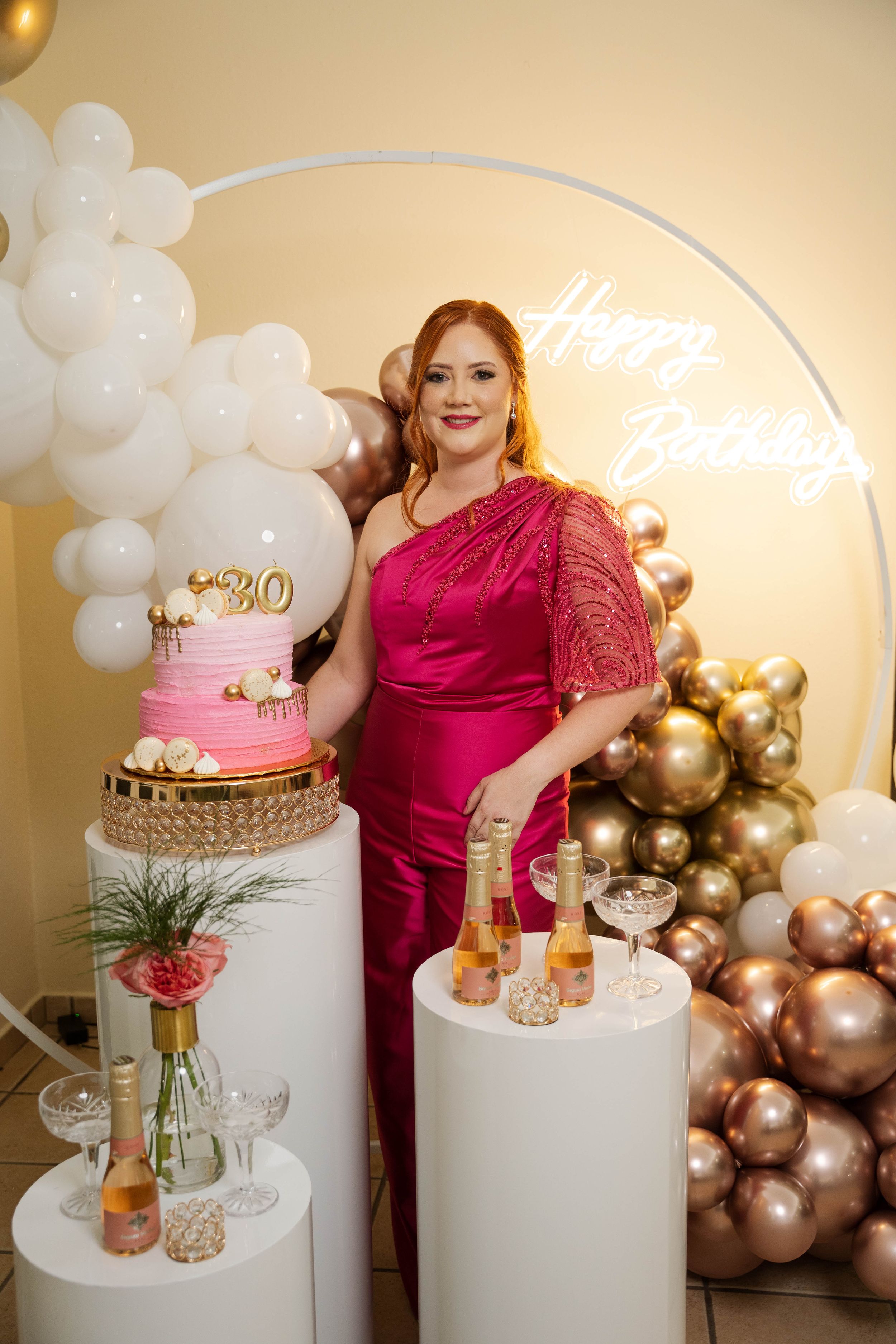 30th birthday party setup with pink cake, balloons, and champagne on white pedestals.