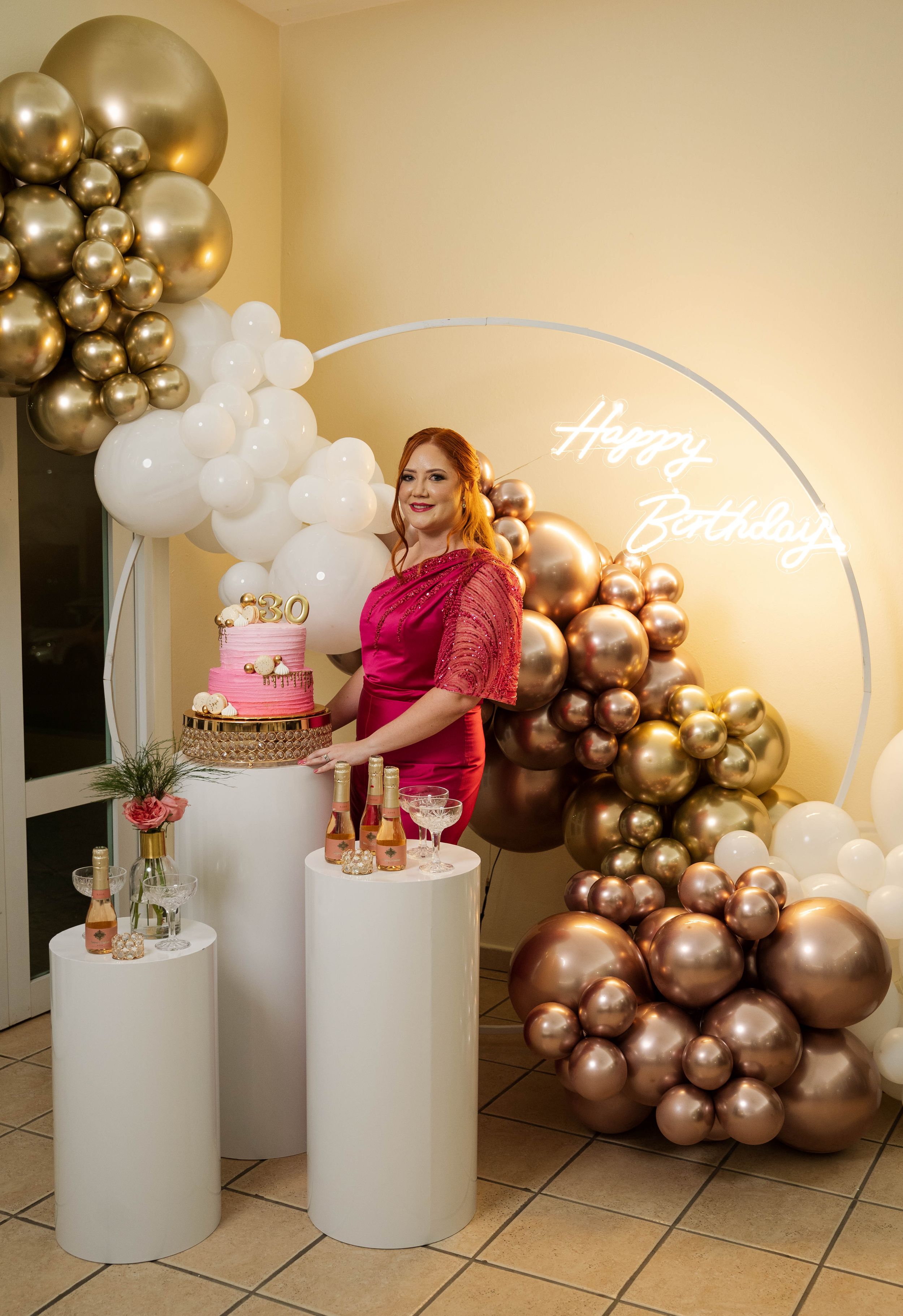 Pink and gold tiered birthday cake displayed on white pedestal with balloon arch and neon sign backdrop.