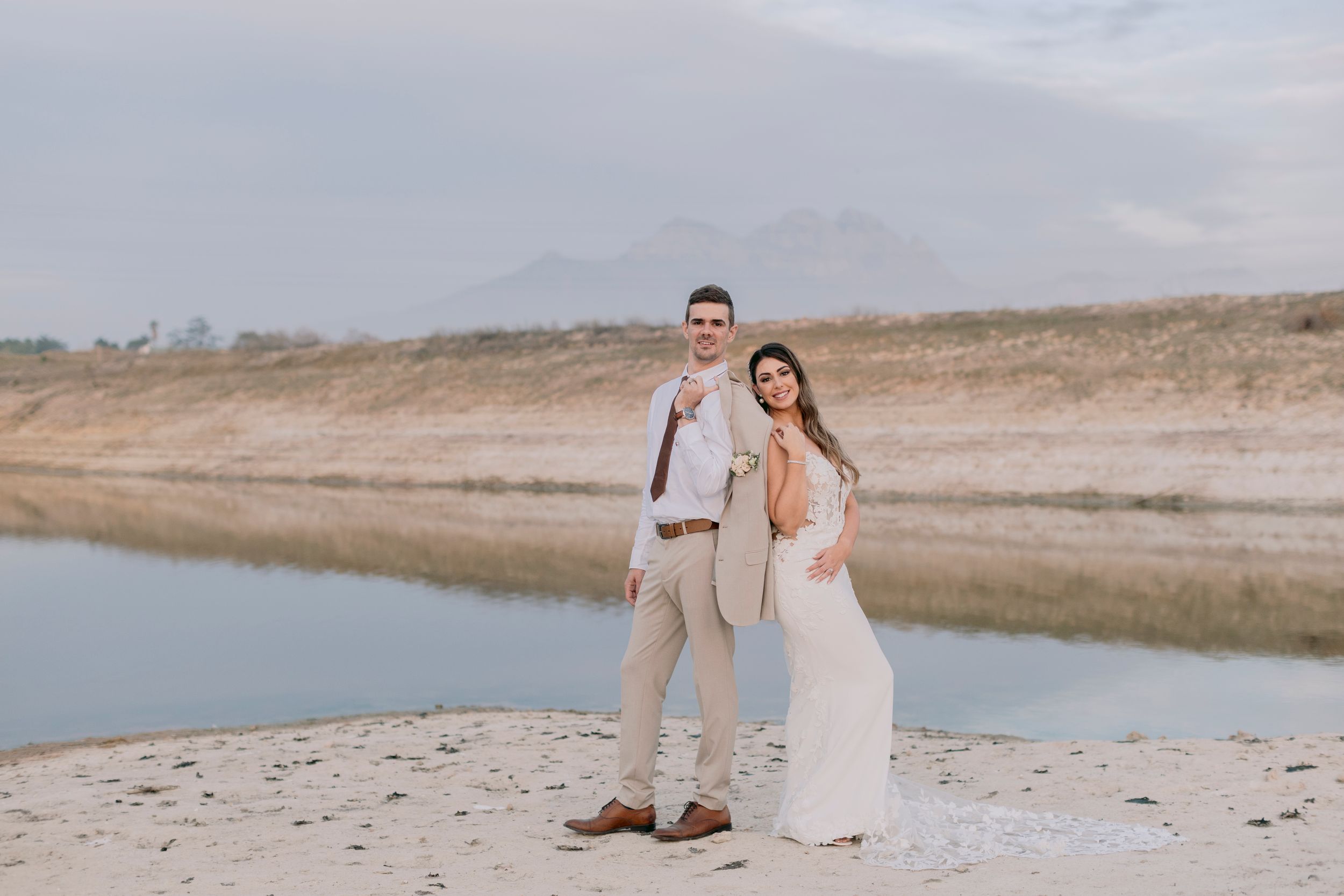 Couple in wedding attire posing on sandy beach with mountain and lake backdrop.