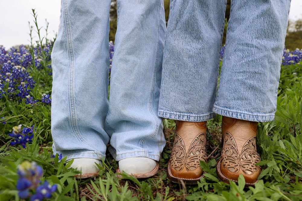 Two pairs of feet in jeans standing among blooming bluebonnet wildflowers.