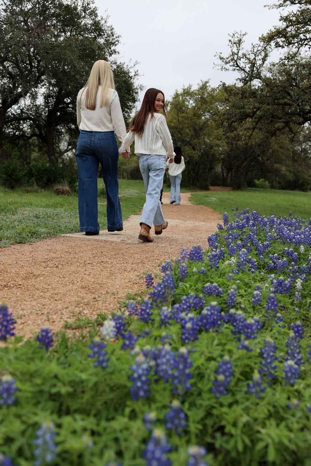 Friends walking on a dirt path bordered by blooming bluebonnets on a cloudy day.