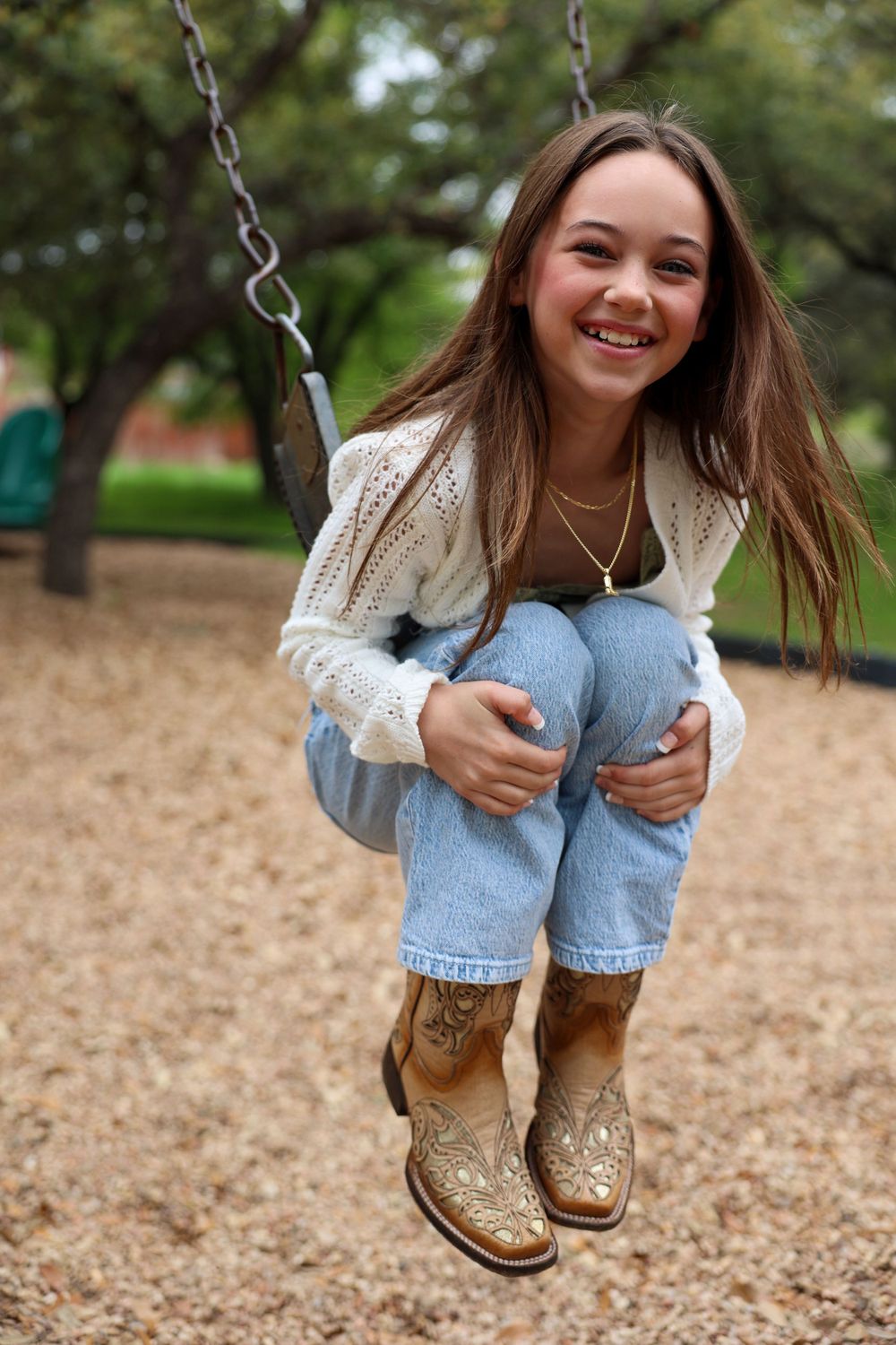 Person on a playground swing wearing a white sweater, jeans, and decorative boots.