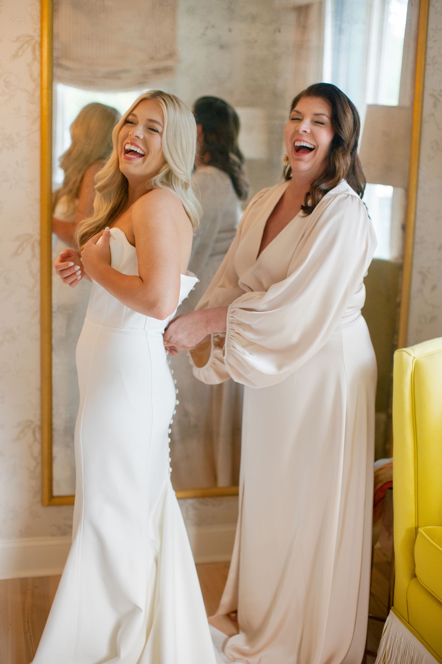 Two women in white dresses share a joyful moment in front of a mirror.