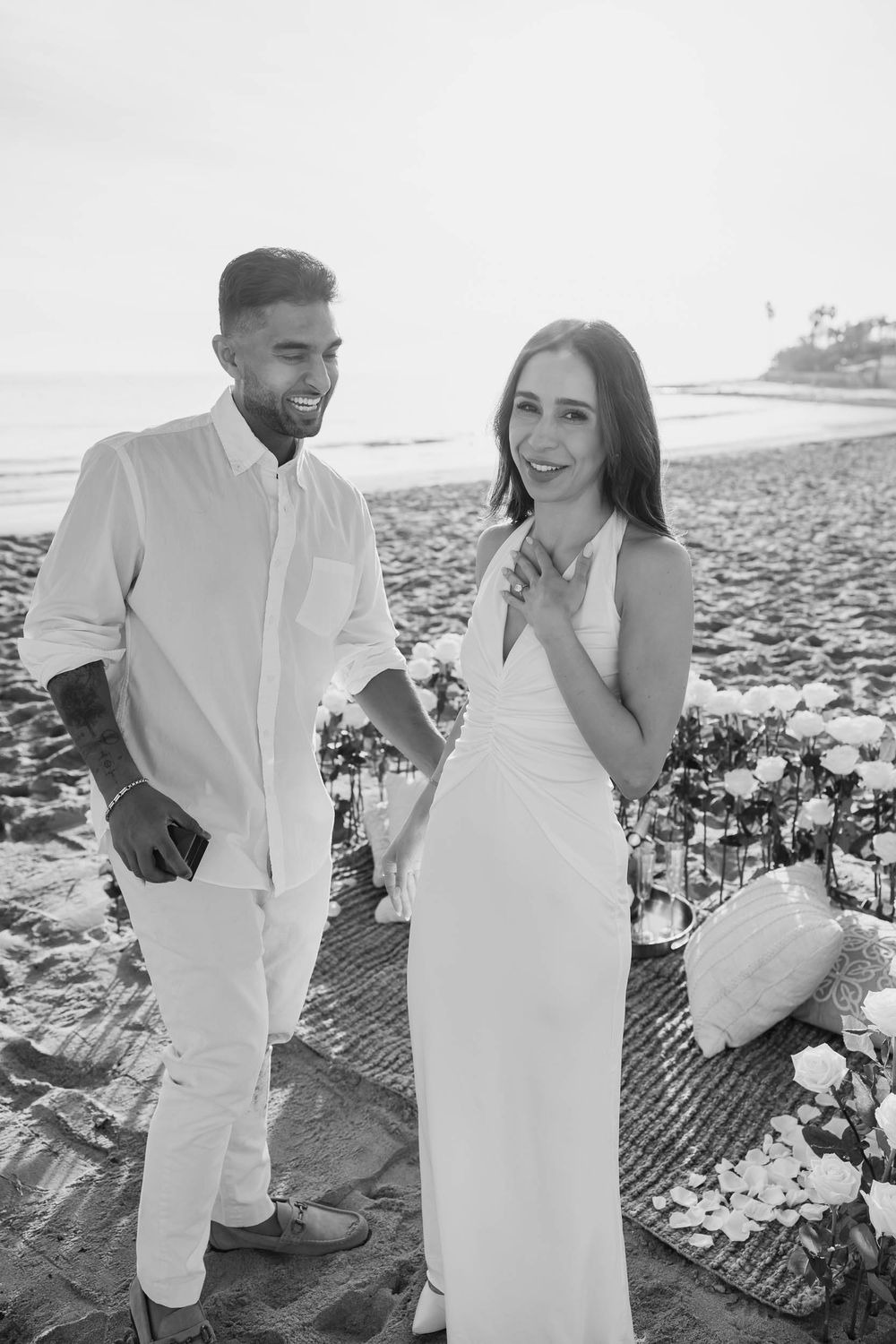 Romantic beach proposal setup with white floral arrangements on sandy shore.