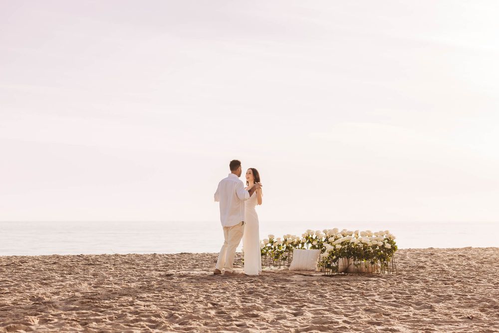 Couple walking along beach with floral arrangements in distance at sunset.