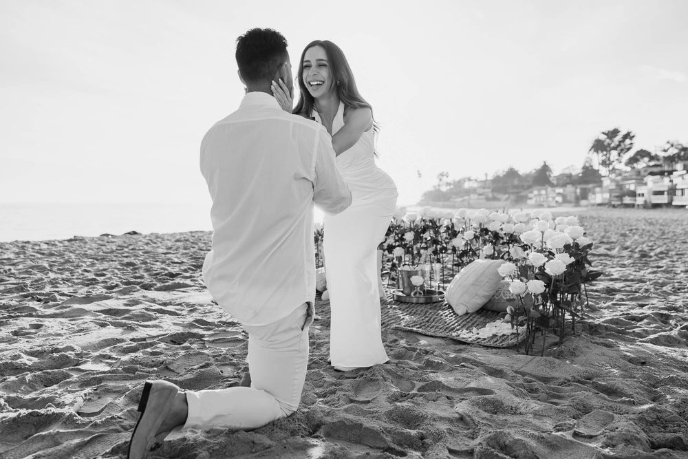 A black and white beach proposal photograph with floral arrangements in the sand.