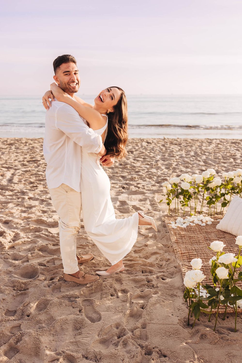 A joyful couple embracing on the beach with white floral decorations.
