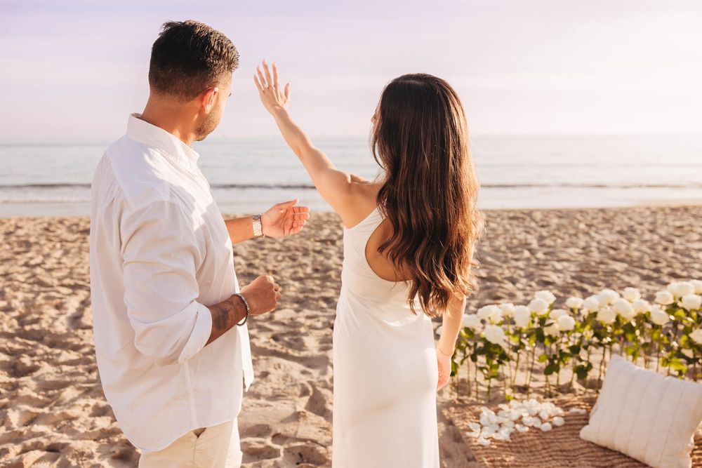 Couple in white outfits sharing a tender moment on a sandy beach during golden hour.
