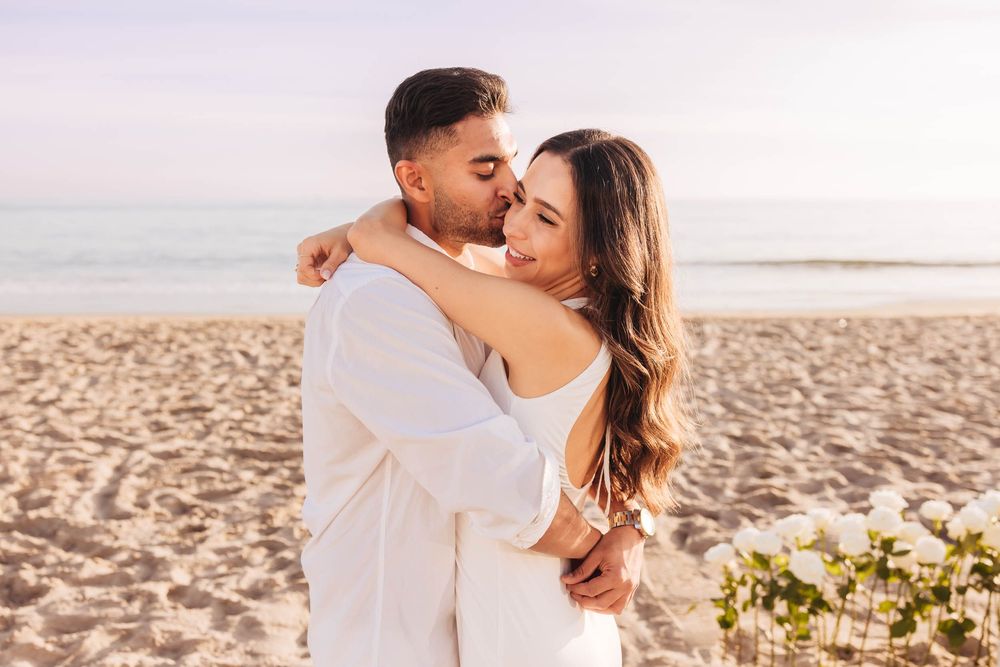 Intimate beach photoshoot of a couple in white clothing embracing during sunset.