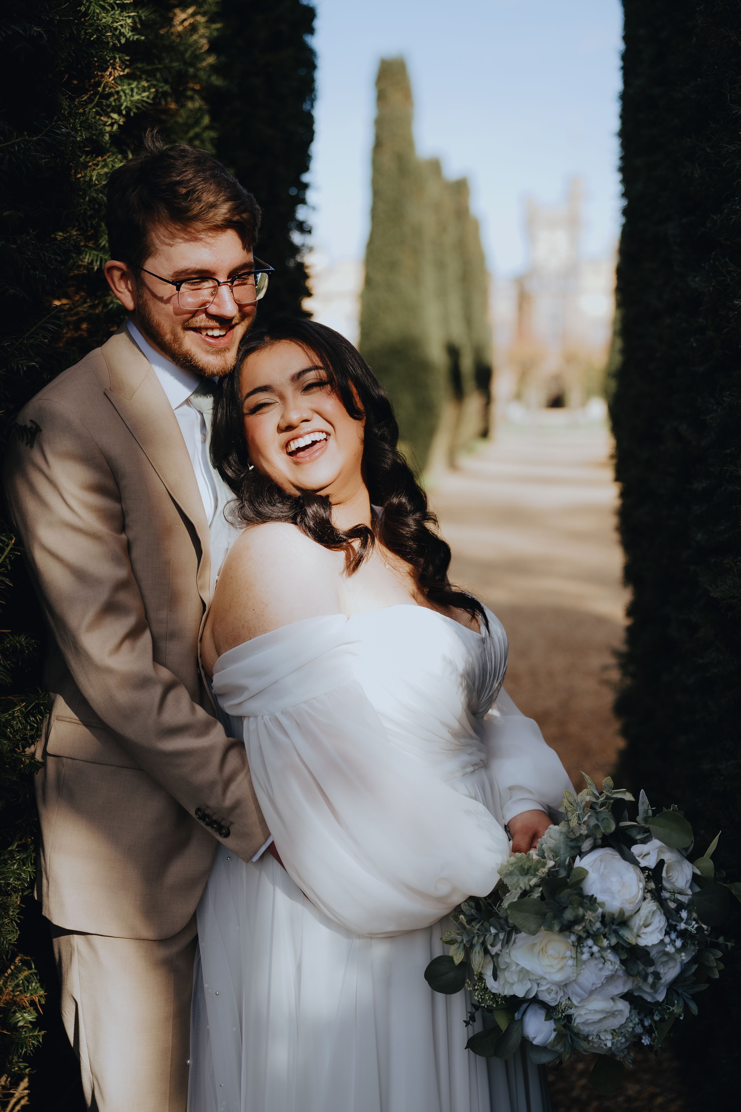 A joyful wedding couple shares a candid moment in a garden path lined with cypress trees, bride holding white bouquet.