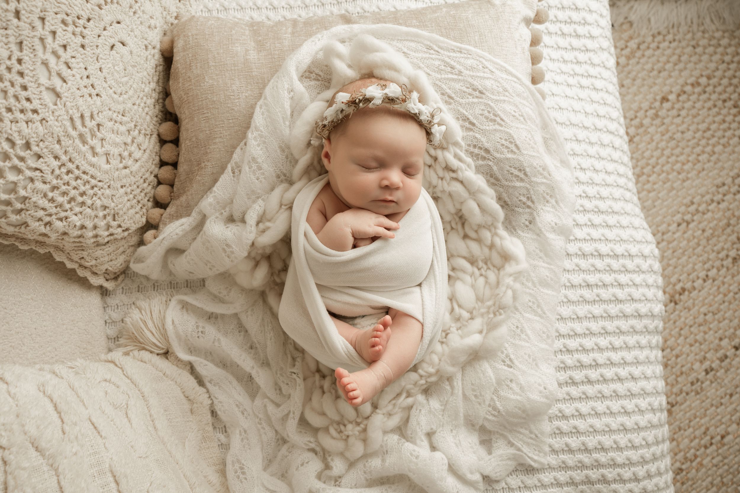 Newborn baby swaddled in white fabric lying on textured cream blanket with delicate lace details.
