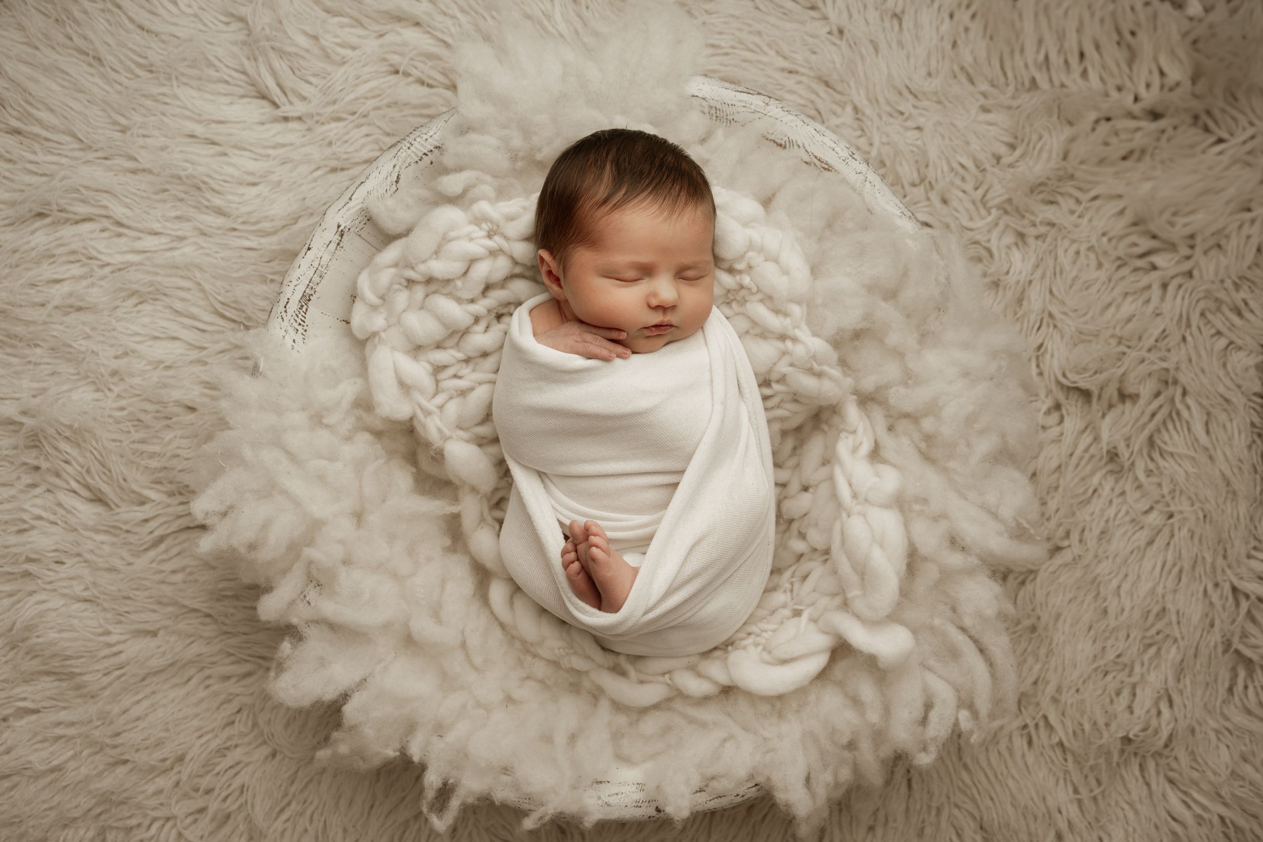 A newborn baby swaddled in white fabric lies peacefully in a textured cream-colored nest-like setting.
