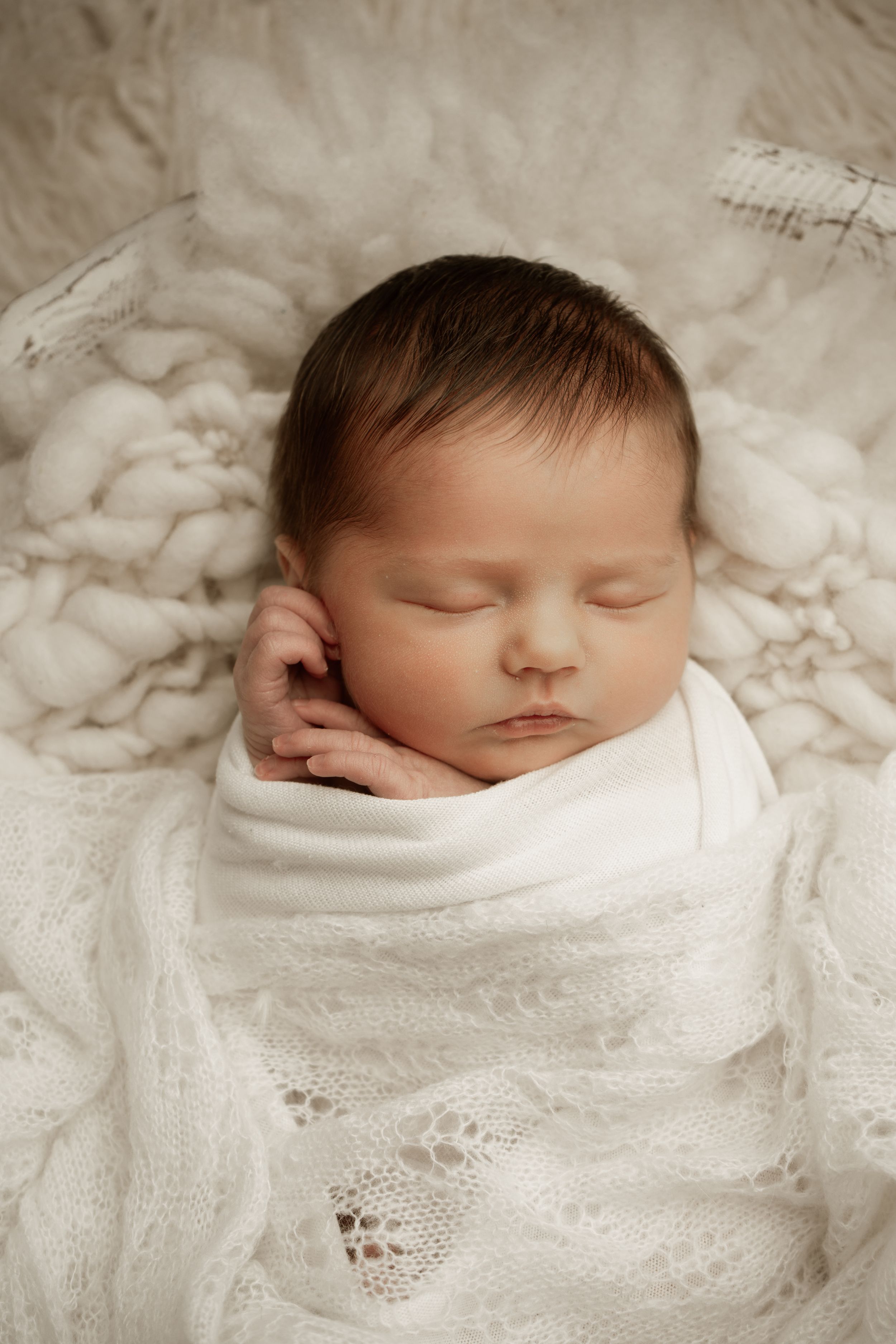 Sleeping newborn baby swaddled in soft white lace blanket rests peacefully on cream textured backdrop.