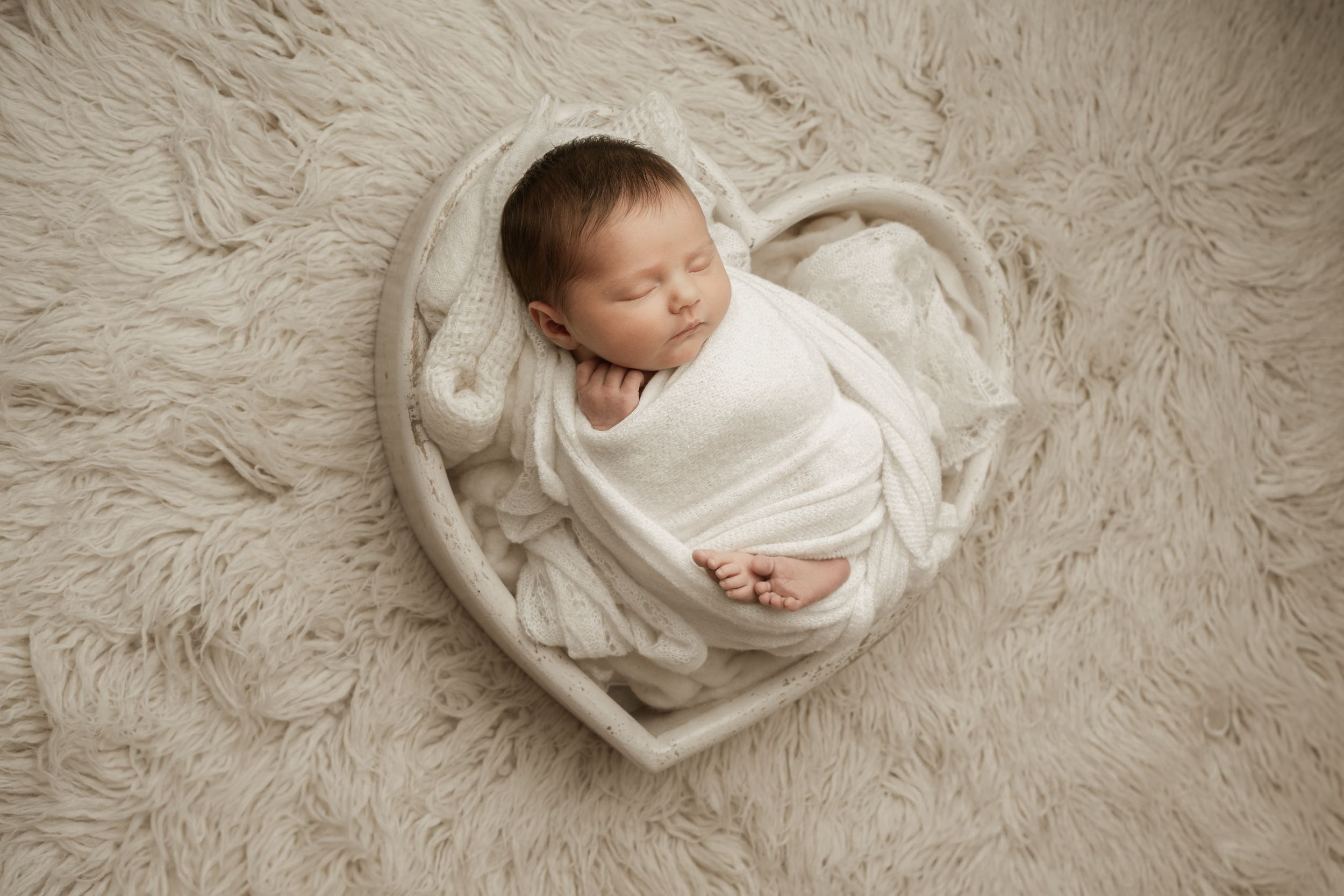 A sleeping newborn wrapped in soft white fabric lies on a textured beige fur blanket in a peaceful pose.
