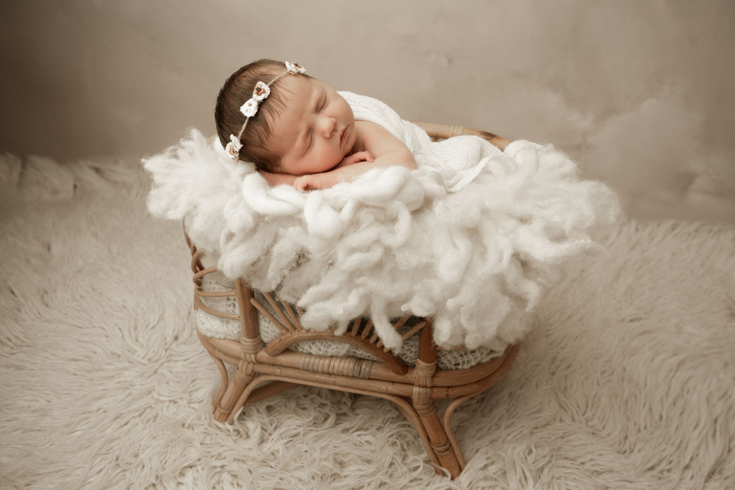 A baby dressed in white rests peacefully on a soft white fur blanket while sleeping in a wooden basket.