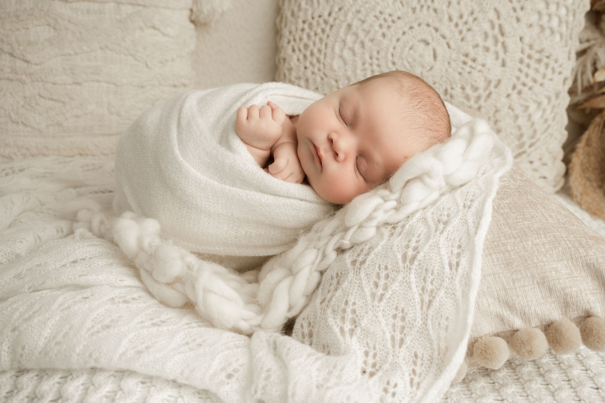 A sleeping baby in a white christening gown lies peacefully on soft white bedding.