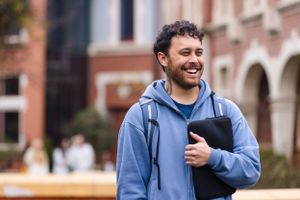 Student wearing blue hoodie carrying laptop and backpack smiling on college campus.