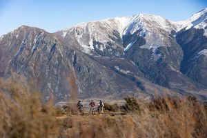 Three mountain bikers ride through golden brush with snow-capped peaks rising dramatically against a clear blue sky.