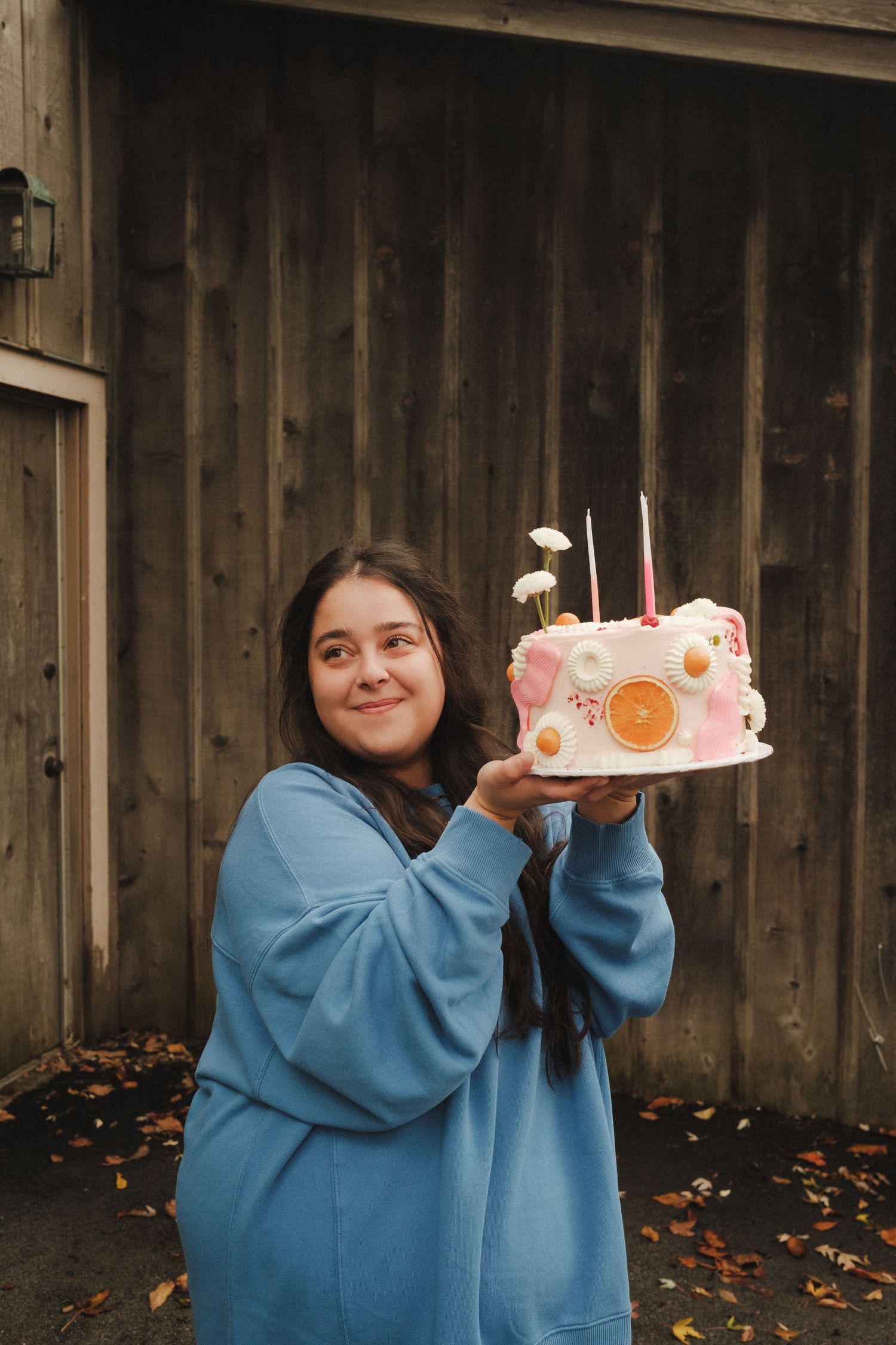 Person in blue hoodie holding up a decorated cake in front of wooden wall.