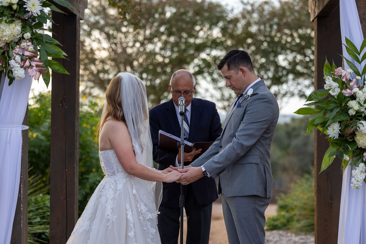 An outdoor wedding ceremony under a decorated arch with white fabric draping.